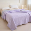 A purple quilted blanket is spread on a bed in a bright room.