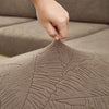 A hand is touching a textured, brown jacquard elastic sofa cushion cover, showing the intricate leaf pattern.