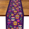 A purple linen table runner featuring a repeating pattern of colorful sugar skulls and floral designs.
