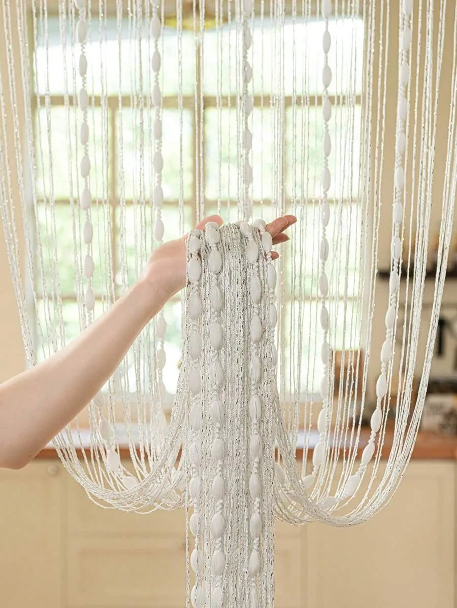 A hand is holding a long, white beaded string curtain with draped fabric, hanging in front of a window.