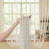 A hand is holding a long, white beaded string curtain with draped fabric, hanging in front of a window.
