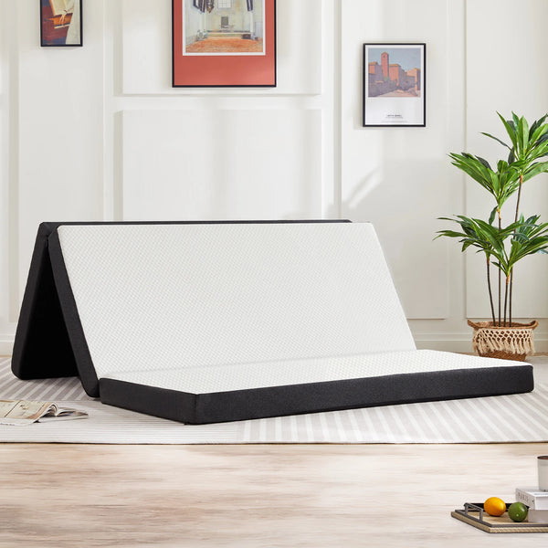 A tri-fold memory foam mattress is shown unfolded on a light wooden floor in a well-lit room.