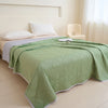 A light green quilted blanket is spread on a bed in a bright room with white bedding and a cream-colored chair.