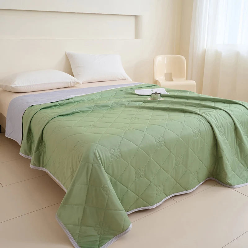 A light green quilted blanket is spread on a bed in a bright room with white bedding and a cream-colored chair.