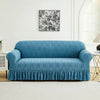 A teal blue sofa cover with a ruffled skirt is displayed against a white wall with a framed floral print.