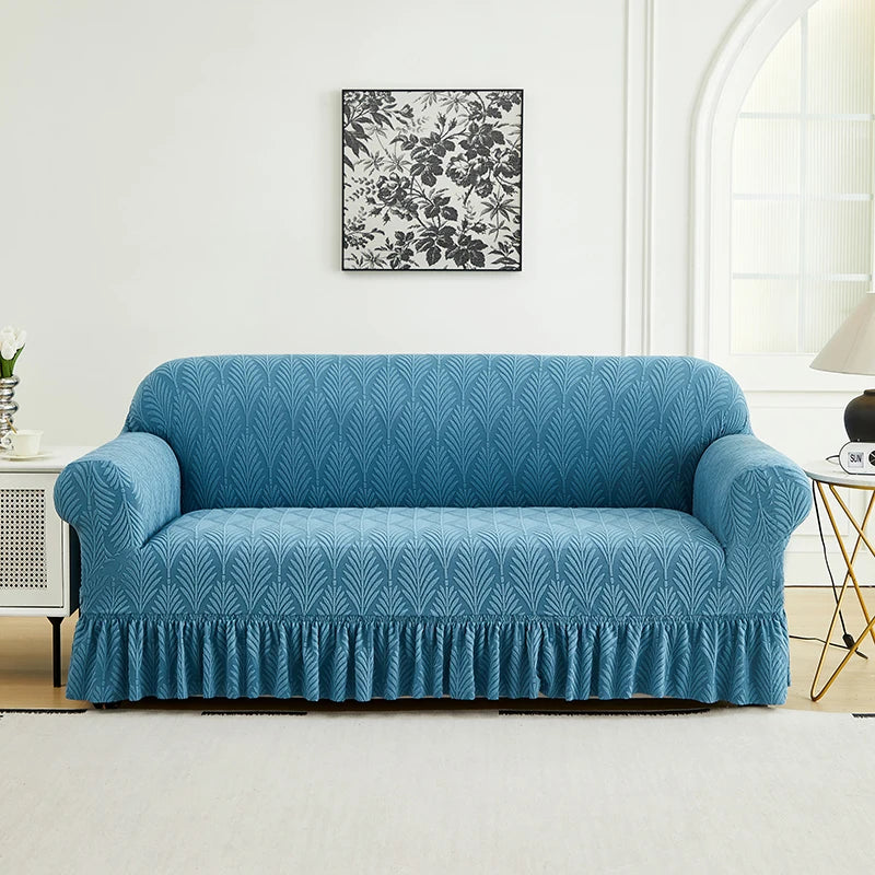 A teal blue sofa cover with a ruffled skirt is displayed against a white wall with a framed floral print.