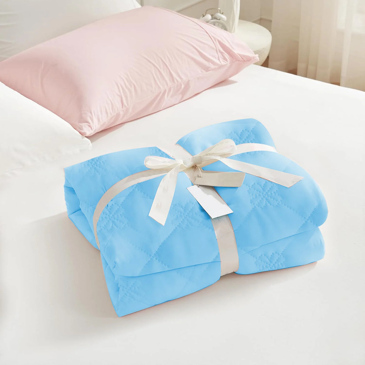 Light blue quilted blanket tied with a white ribbon resting on a white bed.