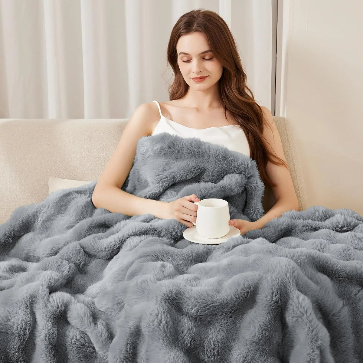 A woman is relaxing on a sofa covered with a gray faux fur rabbit blanket.