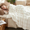 A woman is relaxing under an oversized cream faux fur blanket on a sofa.