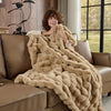A woman is wrapped in a thick, beige faux fur rabbit blanket while sitting on a brown leather sofa.