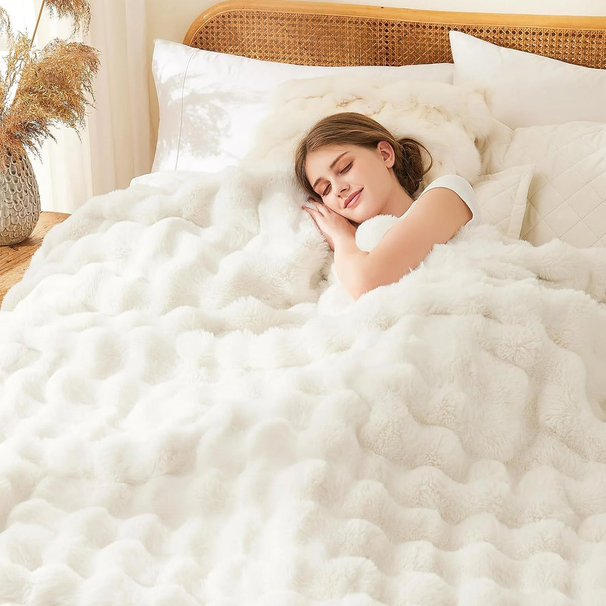 A young woman is relaxing under an oversized white faux fur blanket on a bed.