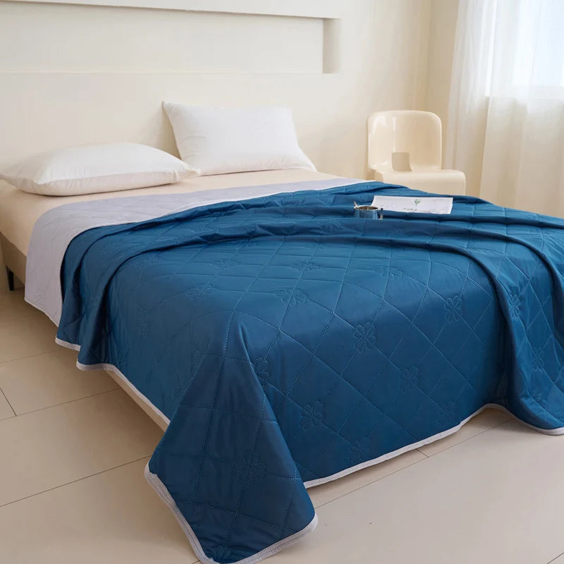 A deep blue quilted blanket is spread on a white bed with white pillows in a bright room.