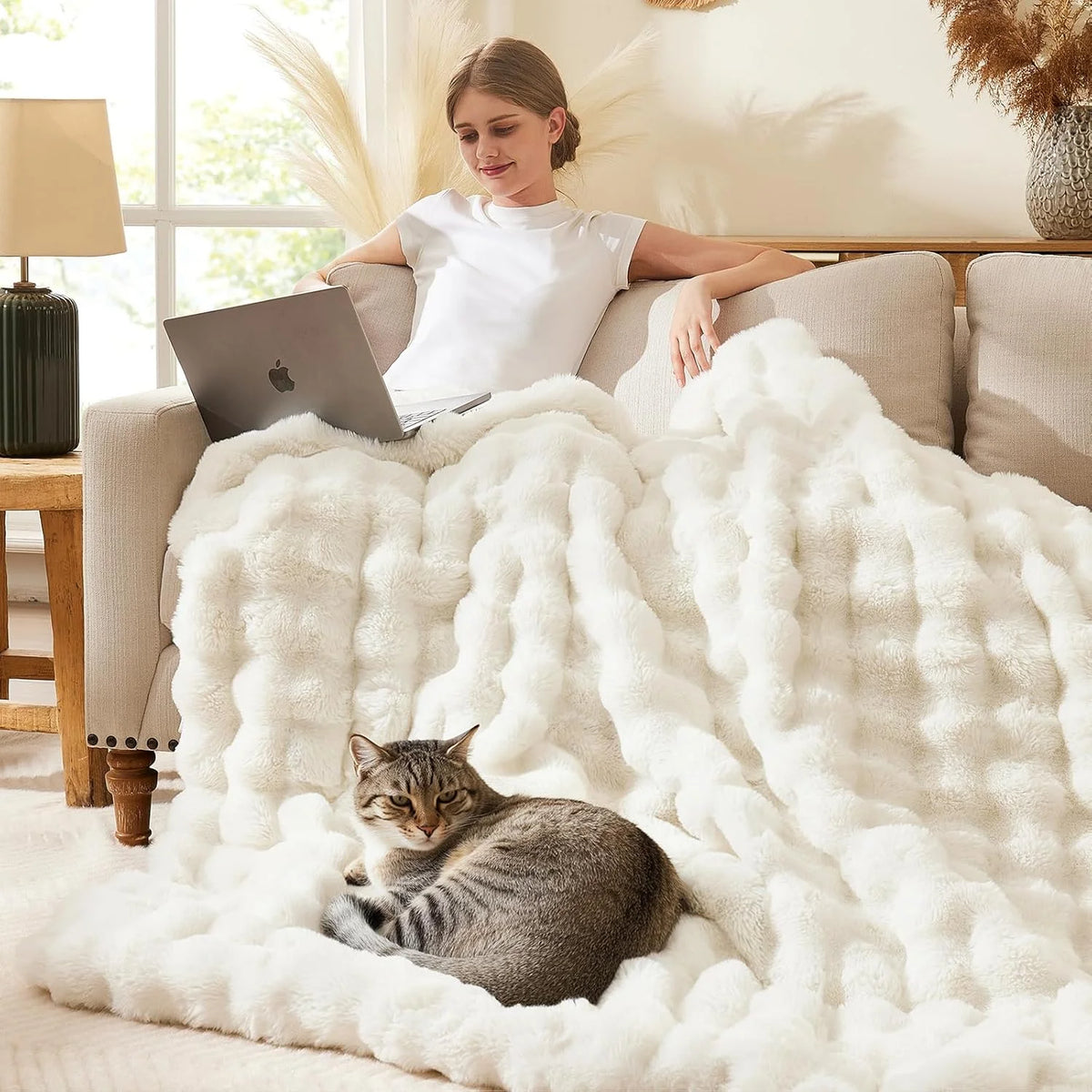 A tabby cat rests on a fluffy white faux fur rabbit blanket next to a woman reading on a sofa.