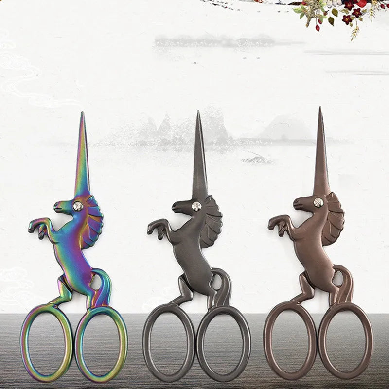 Three stainless steel unicorn sewing scissors with iridescent finishes displayed on a wooden surface.