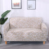 A beige stretch floral elastic sofa cover displayed on a light gray floor with framed art on the white wall.