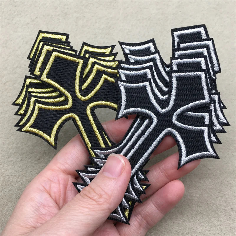 A hand holds several black and gold embroidered cross applique patches for clothing.