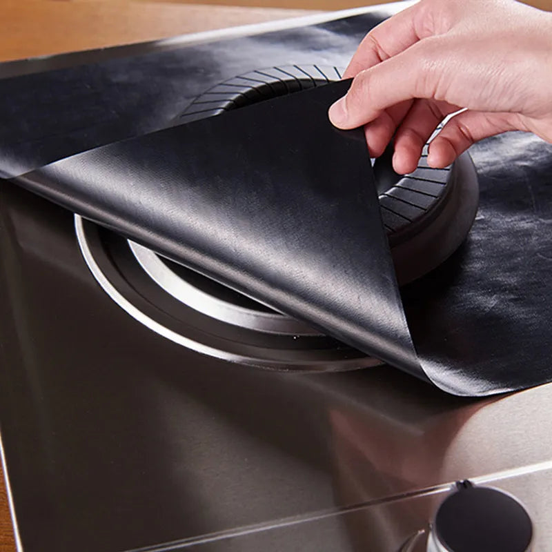 A hand is placing a black reusable non-stick gas stove cleaning mat onto a stovetop burner.