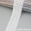A close-up of white cotton embroidered lace trim with a scalloped edge, shown against a gray wooden surface.