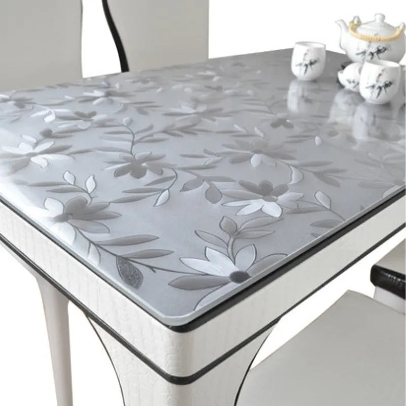 A glass table covered with a silver floral patterned PVC tablecloth and white ceramic cups.
