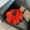 An orange pumpkin shaped plush cushion with black polka dots resting on a black leather sofa.