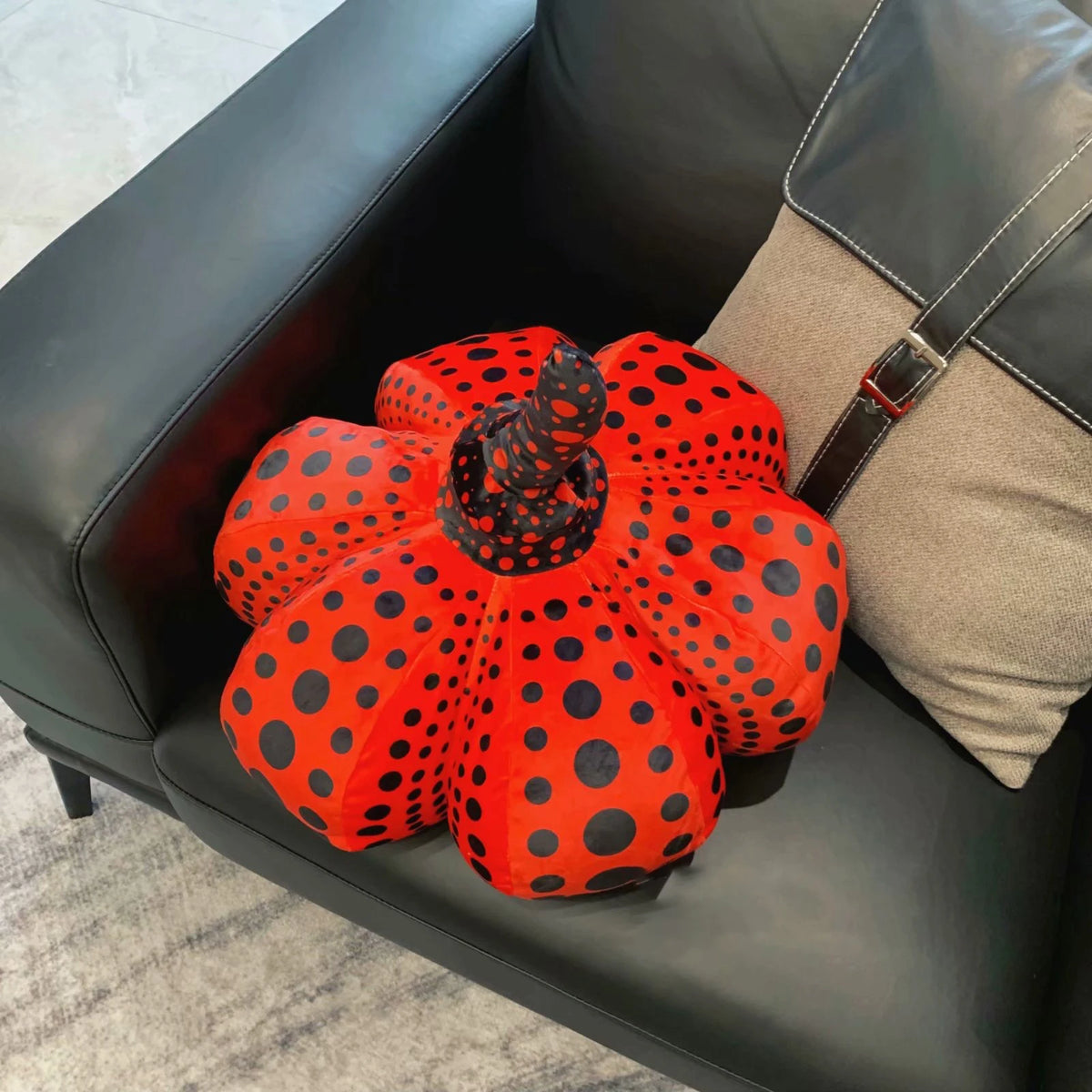 An orange pumpkin shaped plush cushion with black polka dots resting on a black leather sofa.