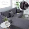 A gray L-shaped sofa protector cover is displayed on a sofa, with a photo frame and a laptop nearby.