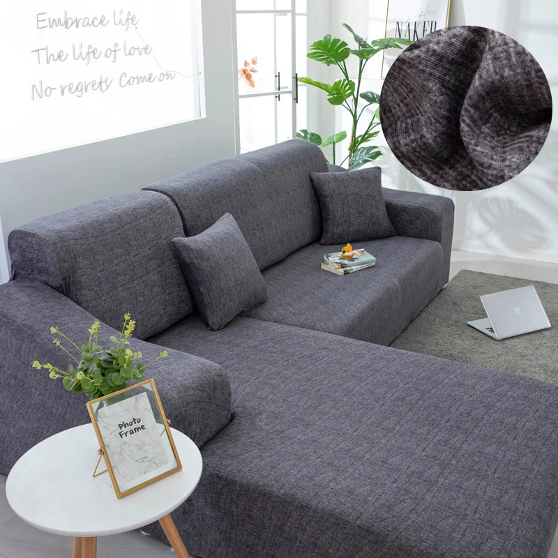 A gray L-shaped sofa protector cover is displayed on a sofa, with a photo frame and a laptop nearby.