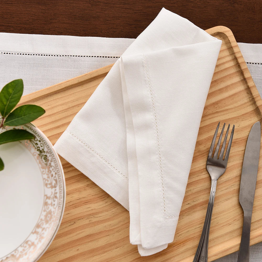 A stack of white linen napkins is placed on a wooden cutting board next to a plate and silverware.