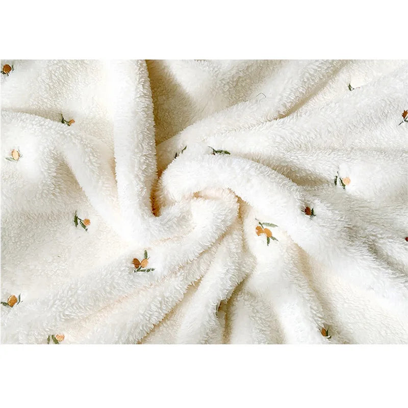 A close-up of a soft, cream-colored coral fleece flannel blanket with subtle green and orange floral embroidery.