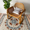 A woven wooden chair with a cushion sits on a patterned rug, next to a green plant.