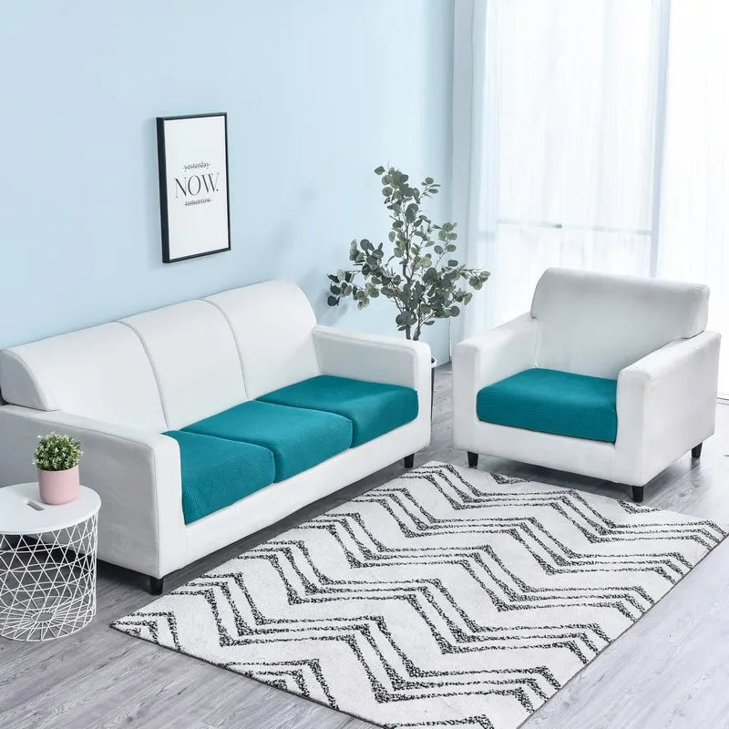 A white stretch sofa seat cushion protector slipcover with teal cushions on a light blue wall.
