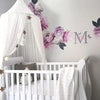 A white crib decorated with a pink floral canopy featuring purple and pink flowers and a large initial M