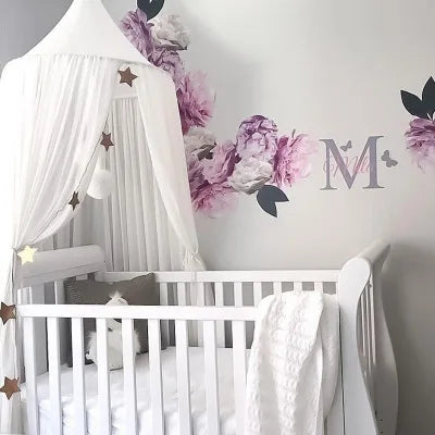 A white crib decorated with a pink floral canopy featuring purple and pink flowers and a large initial M