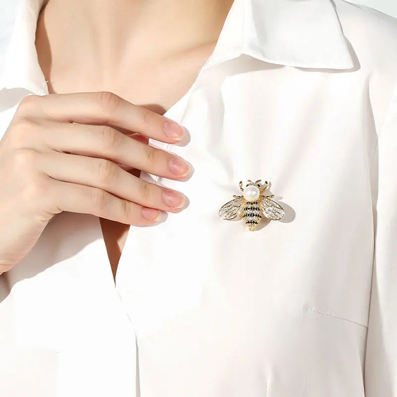 A close-up of a woman wearing a white collared shirt with a crystal bee brooch featuring a pearl accent.