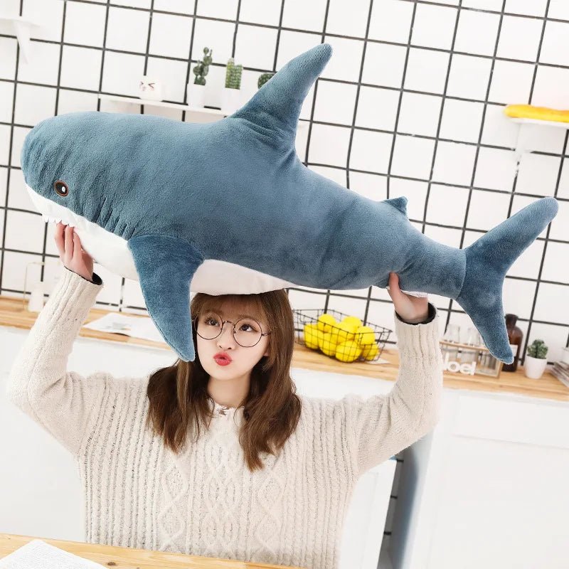 A young woman holds up a large, blue shark plush toy against a white tiled background.