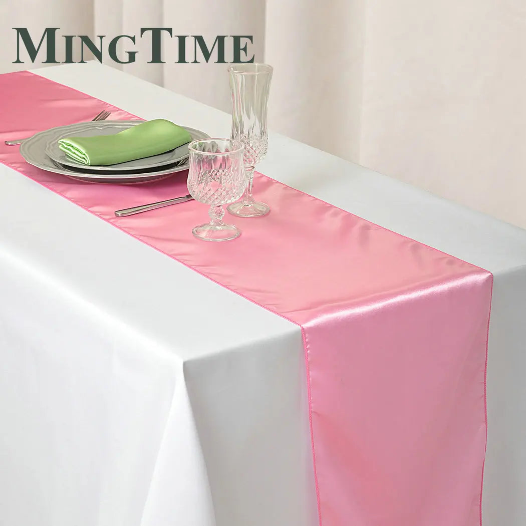 A pink satin table runner is laid on a white tablecloth with silverware and glassware set for an elegant dining experience.