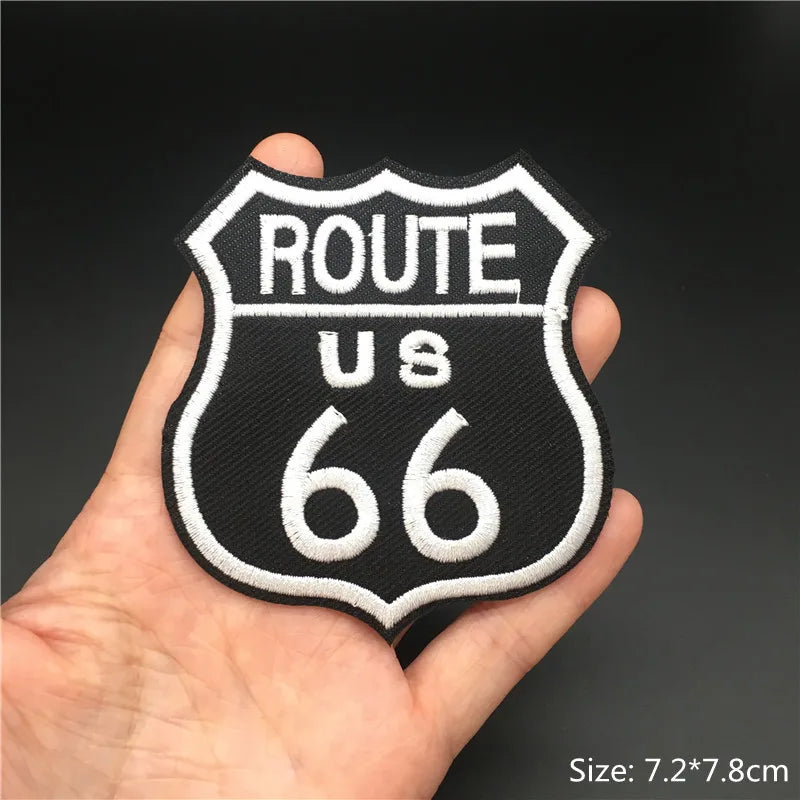 A hand holds an embroidered black and white shield-shaped punk badge with the text ROUTE US 66 on it.
