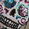 A close-up of a white sequin skull embroidered clothing patch featuring purple, teal, and pink sequins.