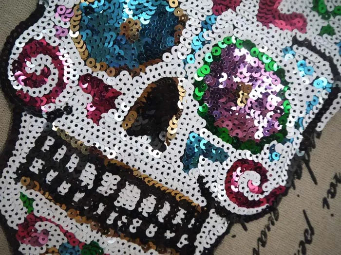 A close-up of a white sequin skull embroidered clothing patch featuring purple, teal, and pink sequins.