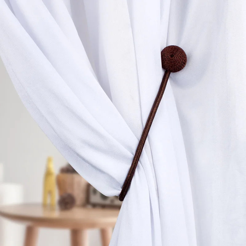 A close-up of a dark brown magnetic pearl ball curtain tieback with a rope handle against a white fabric.