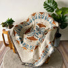 A bohemian geometric throw blanket draped over a wooden chair next to a large green monstera plant.