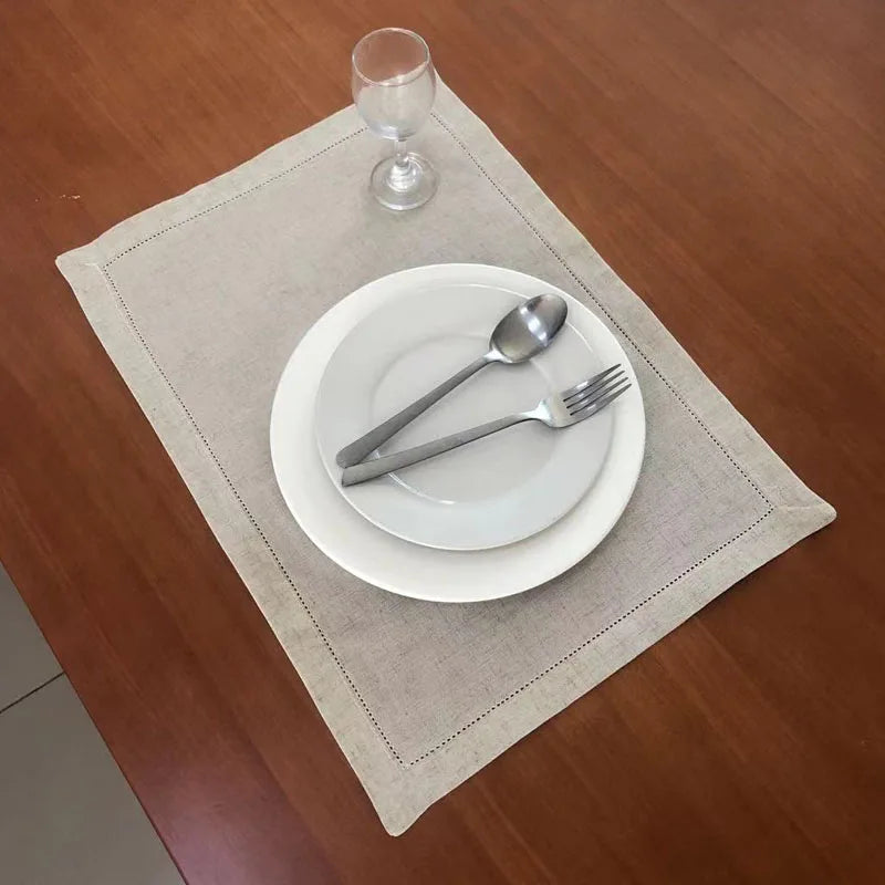 A place setting with a white plate, silverware, and a glass on a beige linen napkin on a wooden table.
