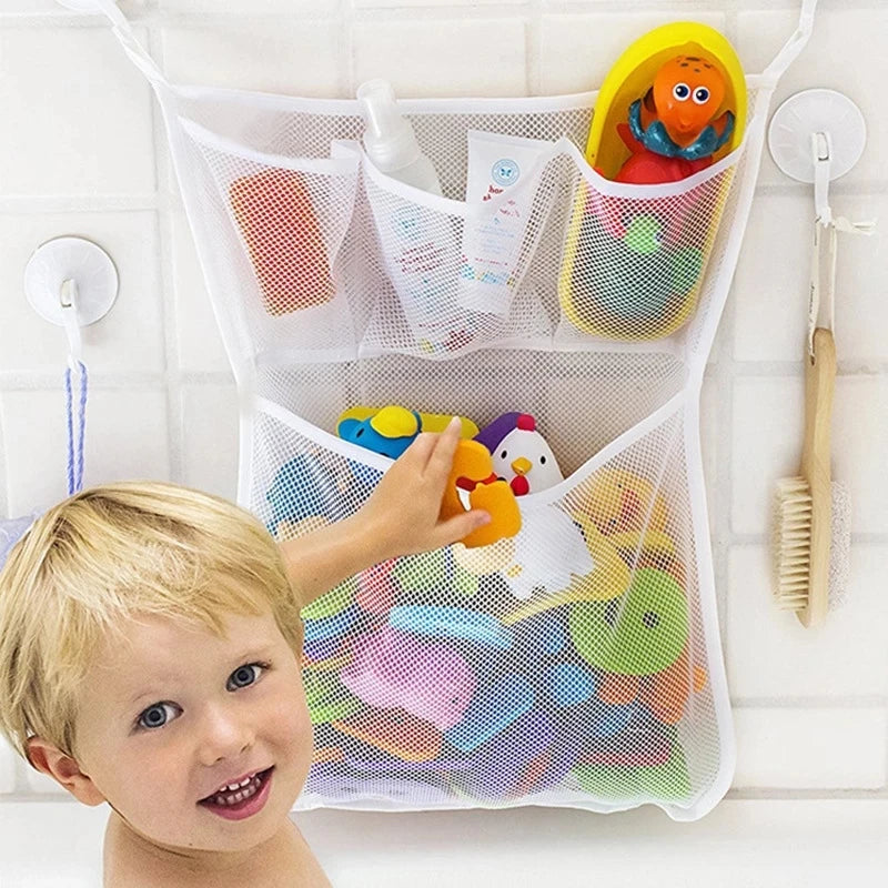 A young boy smiles while reaching into a white mesh storage bag filled with colorful bath toys, including a duck and frog.