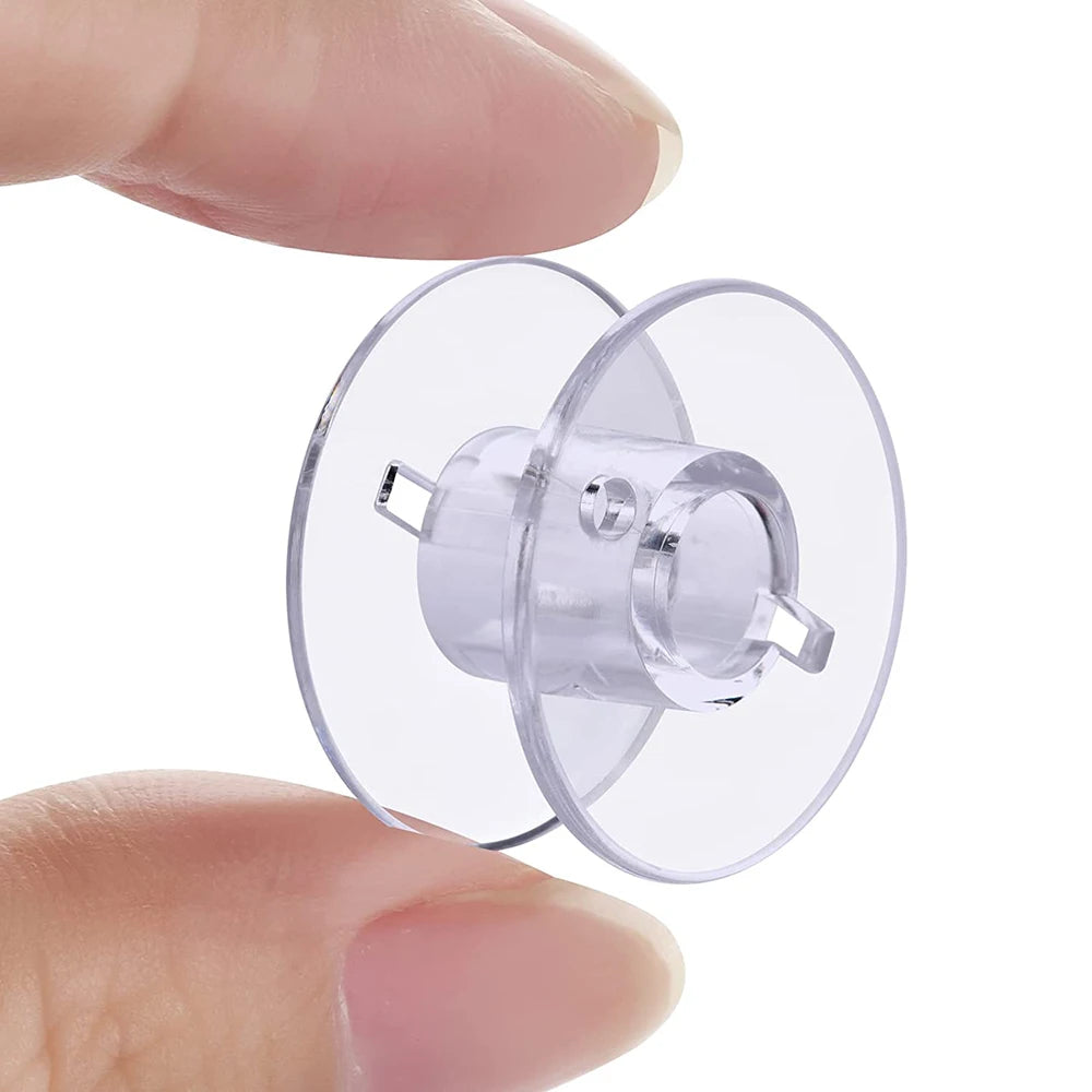 A hand holds a transparent plastic sewing bobbin with a metal holder and a clear plastic spool.