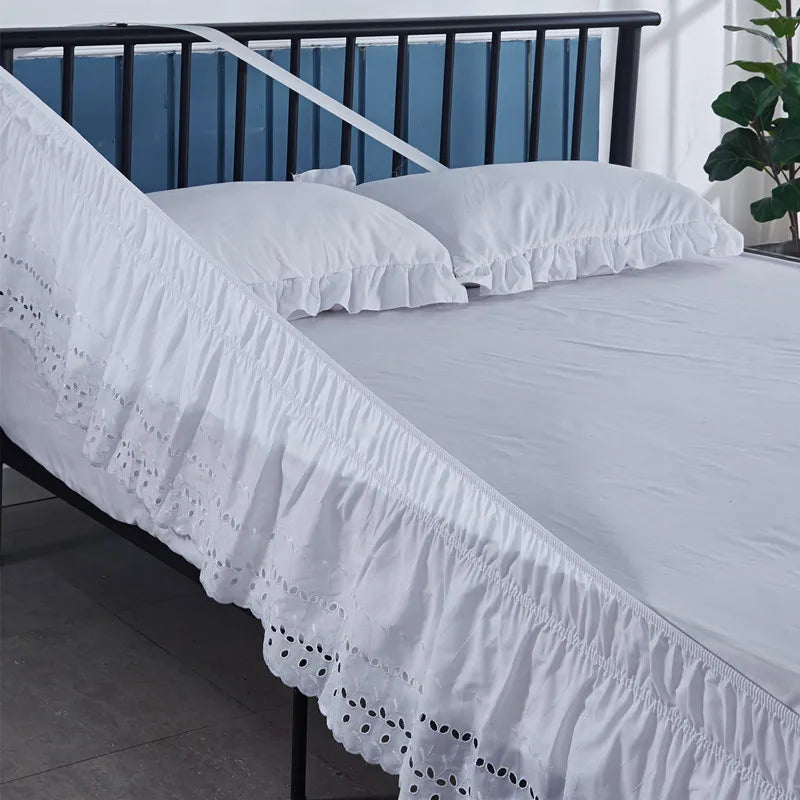 White embroidered ruffle bed skirt with white linens and pillows on a black metal bed frame.