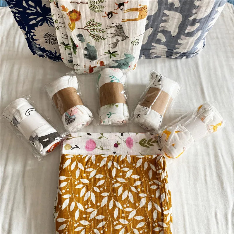 A white muslin swaddle blanket with floral embroidery is displayed on a white bed, surrounded by rolled fabric.