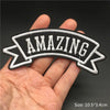 A hand holds an embroidered black and white banner with the word AMAZING on it.