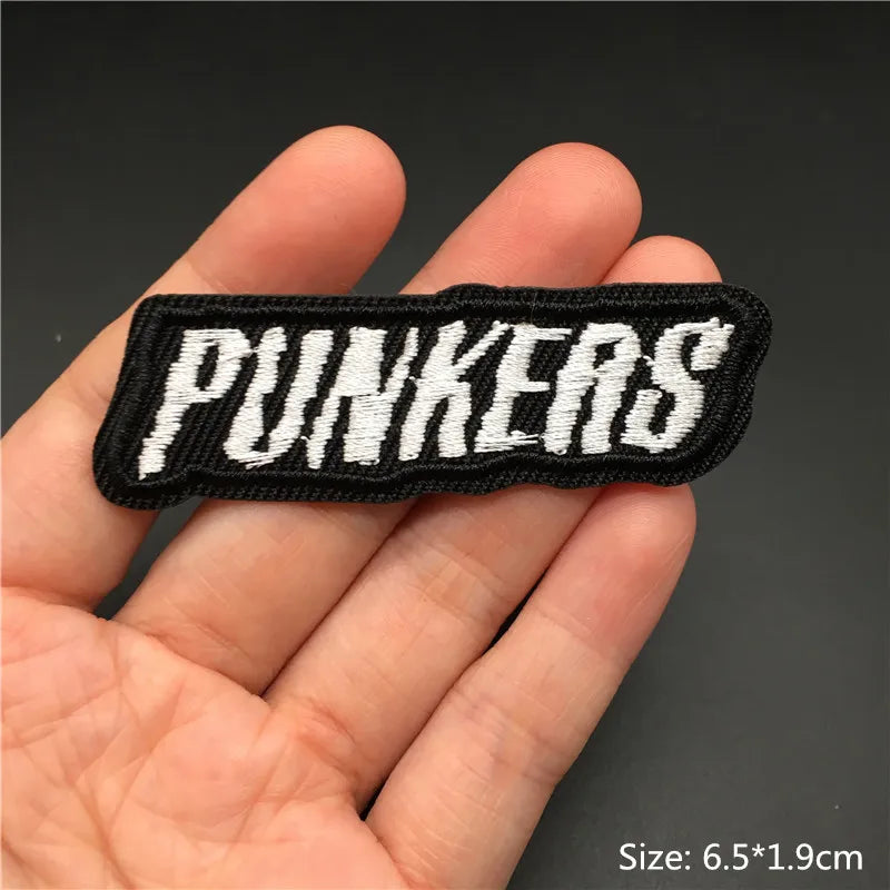 A hand holds a black embroidered punk badge with the word PUNKERS stitched in white thread.
