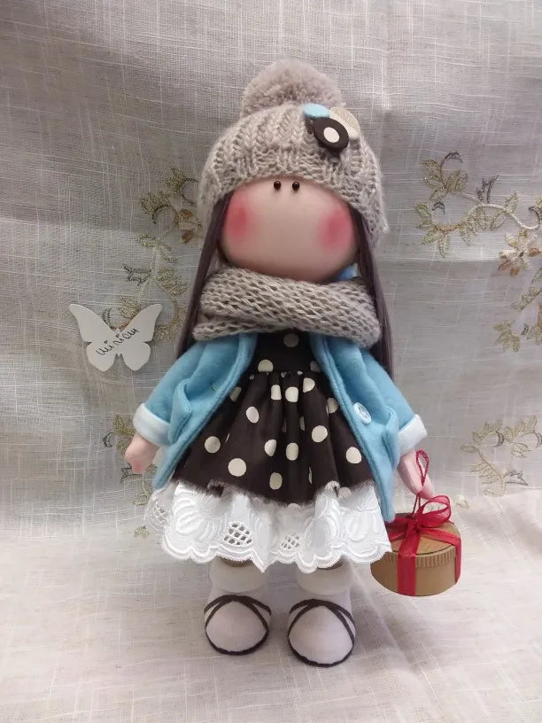 A handmade cotton toy filler batting doll dressed in a gray knitted hat and blue clothing.