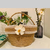 A woven basket made of natural jute rope with a handle, decorated with white lace trim and a large flower.
